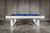 Doc & Holliday Santorini Pool Table, a sleek white pool table with a blue playing surface, set against a wooden wall with a concrete floor.