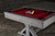 Doc & Holliday Hepburn Pool Table, a stylish modern pool table with a red felt playing surface and a set of billiard balls arranged on it, against a dark brick wall background.