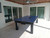 Sawyer Twain Metro All Weather Outdoor Pool Table with Storage Drawer, a rugged blue ping pong table with black legs positioned on an outdoor patio under a wooden ceiling with a ceiling fan, large sliding glass doors in the background.