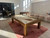 Brunswick Billiards Brunswick Parsons Pool Table in Natural Dry Oak, in an indoor game room setting with sunlight, pool cues on the table, and bar stools against a textured wall.