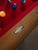 Doc & Holliday Hank Slate Pool Table – Americana Chocolate, a close-up of the wooden edge and the metallic brand plate on the pool table with balls on the red felt surface.