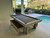 American Heritage Billiards Lanai Outdoor Slate Pool Table in Oyster Grey, pool table on indoor patio with screened enclosure, nearby pool and outdoor seating.