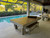American Heritage Billiards Lanai Outdoor Slate Pool Table in Oyster Grey, located in an outdoor patio with seating area and pool, showcasing its sleek design and durable materials.