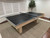 American Heritage Billiards Vancouver 8' Slate Pool Table in Natural Ash, featuring a sleek black playing surface with a netted divider, situated in a modern room with white walls, plantation shutters, and a potted plant.