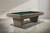 Three-quarter angle view from a professional photo shoot of the Doc & Holliday Preakness slate pool table, featuring a glazed pearl wood finish, solid slate construction, Championship green felt, wide rails, and tapered mid-century modern block legs.