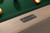 Doc & Holliday Preakness Pool Table in Glazed Pearl, close-up of the wooden frame with brand metal plaque, green felt surface, and billiard balls.