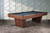 Doc & Holliday Preakness Pool Table in Gray Walnut, modern billiards table with a sleek wooden finish and blue felt surface, set against a contemporary wall with geometric patterns.