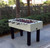Sawyer Twain Outdoor Foosball, a white outdoor foosball table with black legs and red and black players, set up on a paved area outdoors with greenery in the background.