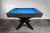 Wide lifestyle view of the Jett Stealth Edition showing its full steel leg design, slim aprons, and Smokehouse maple rails. The bold blue felt draws the eye while the sleek proportions make a strong impression across open interiors, hospitality lounges, or modern game rooms.