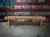 Doc & Holliday Hank Shuffleboard Table in Oaklee finish staged in front of a rustic American flag backdrop. Features solid oak frame, recessed scoring zones, and finger-jointed aprons, set on an industrial concrete floor.