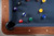 Doc & Holliday Preakness Slate Pool Table in Walnut & Maple, featuring a blue felt surface with billiard balls arranged on it, surrounded by a wooden frame with decorative inlays.