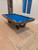 Contemporary Essex Pool Table in all-steel gunmetal construction with precision-cut base legs and blue billiard cloth. A designer centerpiece for modern game rooms, shown on light maple flooring beside dining area.