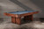 Doc & Holliday The Jackies Slate Pool Table, featuring a Bourbon Pecan Walnut finish with sleek slate surface, set against a gray wooden wall.