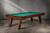 Doc & Holliday Muse Slate Pool Table by Doc & Holliday, a wooden pool table with green felt surface and angled legs, set against a gray wooden-paneled wall.