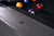 American Heritage Billiards Alta Pool Table in Marble Grey, close-up of the textured wood surface with a black diamond inlay, and billiard balls in the background.