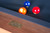 American Heritage Billiards Vancouver Slate Pool Table in Honey Ash, close-up of the wooden frame with a metal plaque and the blurred pool balls in the background.