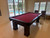 Brunswick Billiards Brunswick Oakland 8' Slate Pool Table, espresso finish, with red felt, located indoors near large windows and a sliding glass door overlooking a backyard pool.
