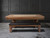Doc & Holliday Kaci Slate Pool Table – Glazed Maple, a wooden pool table with a natural glazed maple finish and a black padded bench, set against a dark textured wall.