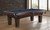 C.L. Bailey Adrian Slate Pool Table, a classic wooden pool table with a dark finish and a slate playing surface, situated in a modern room with large windows and a brick wall.