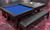 C.L. Bailey Tunbridge Pool Table & Shuffleboard in Weathered Brown, with a blue shuffleboard surface and a dark brown wooden frame, set on a patterned red and black carpet with a dining table and bench nearby.