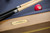 Plank & Hide Co Plank & Hide Deco 8' Slate Pool Table in Glazed Maple, close-up of the wooden table edge with a gold nameplate and pool cue sticks with a red ball in the background.
