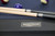 Doc & Holliday Savant 8' Slate Pool Table in Black Ash, close-up of the table's corner with a metal nameplate and pool cues on the table