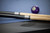 Doc & Holliday Peter Parsons Slate Pool Table in Matte Black Lacquer, pool cues and a billiard ball on a blue felt table with a close-up view of the embossed brand logo.