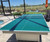 Sawyer Twain Matrix Outdoor Slate Pool Table with Sunbrella Felt, outdoor pool table with blue playing surface and white frame, set up on patio near a lake with outdoor furniture and umbrellas.
