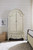 Hooker Furniture Kandor Leather Stationary Sofa, elegant vintage-style white wooden armoire with curved top and brass hardware, placed in a cozy bedroom setting with a woven basket, framed artwork, and a bed with a beige classic bed frame.