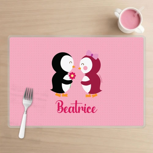 Spark & Spark Penguins in Love Kids Placemat, pink placemat with adorable penguin illustration forming a romantic scene, personalized with the name Beatrice, set on a wooden table alongside a fork and a cup of pink drink.