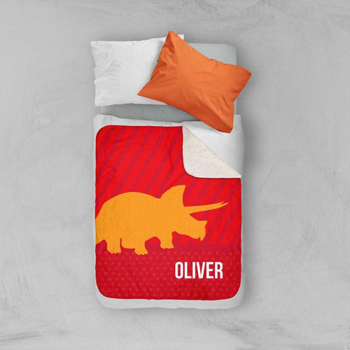 Spark & Spark Dino and Me - Red Sherpa Blanket, red blanket with a yellow triceratops dinosaur silhouette and the name Oliver, placed on a bed with white and orange pillows.