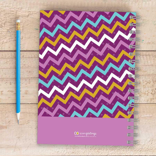 SparkGreetings® Mix Chevrons Writing Journal, spiral-bound notebook with colorful zigzag chevron pattern in purple, blue, white, and gold on a wooden surface with a matching blue and gold pencil beside it.