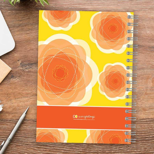 SparkGreetings® Pretty Blooms Writing Journal, a colorful spiral-bound notebook with orange and yellow floral design on the cover placed on a wooden desk.