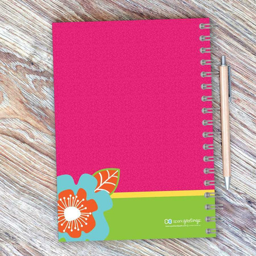 SparkGreetings® Modern Blooms Writing Journal, vibrant pink cover with colorful floral design, spiral binding, and matching pen on wooden surface.