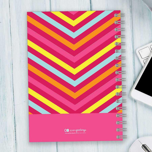 SparkGreetings® Style in Stripes Writing Journal, colorful spiral-bound notebook with a vibrant chevron pattern in pink, yellow, orange, and blue on a wooden surface.