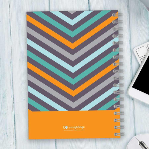 SparkGreetings® Style in Stripes Writing Journal, colorful striped pattern with orange accent at the bottom, spiral-bound notebook on a light wooden desk with a smartphone and keyboard.