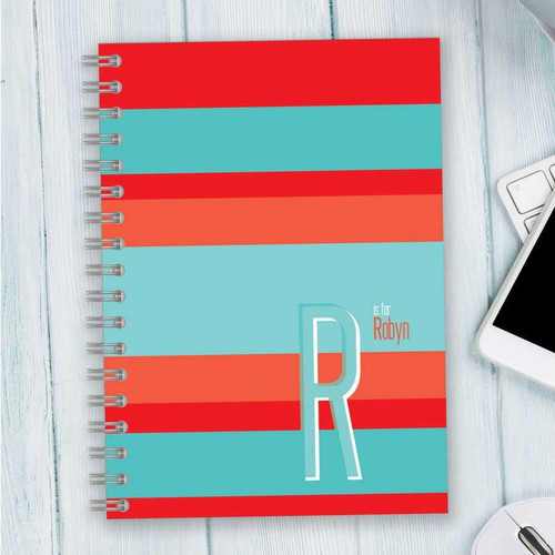 SparkGreetings® Bold and Modern Writing Journal, colorful striped cover with large letter R and personalized name Robyn, on a light wooden surface with part of a laptop and mouse visible.