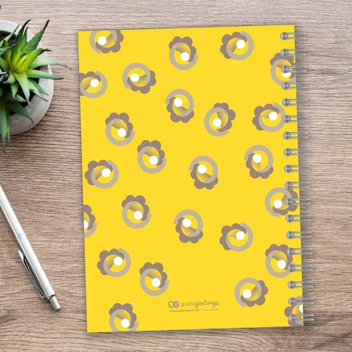 SparkGreetings® Small Swirls Writing Journal, a yellow spiral-bound notebook with a floral swirl pattern on the cover, placed on a wooden desk next to a silver pen and a plant.