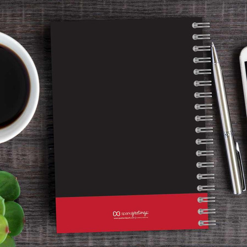 SparkGreetings® Victorian Bold Style Writing Journal, black cover with red bottom band and silver pen on wooden desk with coffee cup and plant.