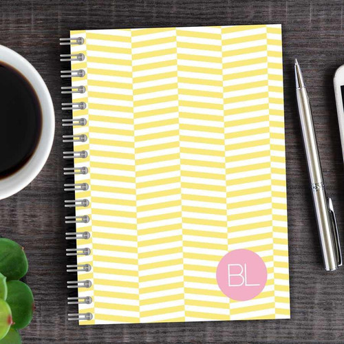 SparkGreetings® Up and Down Style Writing Journal, yellow and white chevron pattern notebook with pink circle and white initials 'BL', on a dark wooden desk with a cup of coffee, a silver pen, and a smartphone.