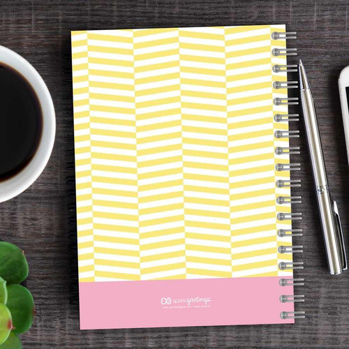 SparkGreetings® Up and Down Style Writing Journal, yellow and pink zigzag patterned notebook with spiral binding on a wooden desk, with a cup of coffee, pen, and smartphone nearby.