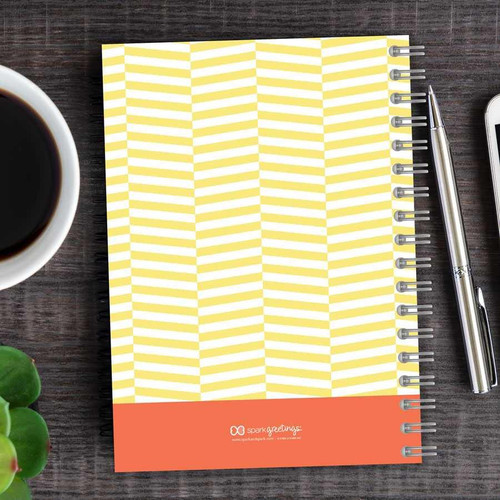 SparkGreetings® Up and Down Style Writing Journal, spiral-bound notebook with yellow and white geometric pattern cover on a wooden desk, next to a silver pen, a cup of coffee, and a smartphone.