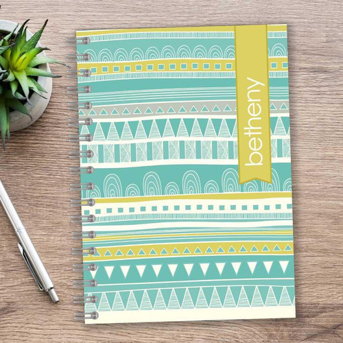 SparkGreetings® Tribal Mood Writing Journal, colorful tribal pattern cover with geometric designs in shades of blue, white, and yellow on a wooden desk.
