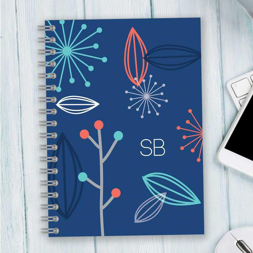 SparkGreetings® Deconstructed Nature Writing Journal, a blue spiral-bound notebook with abstract floral and leaf designs on the cover, placed on a light wooden surface next to a keyboard and a smartphone.