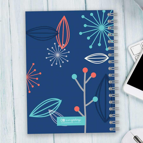 SparkGreetings® Deconstructed Nature Writing Journal, vibrant blue cover with modern abstract floral patterns in red, white, teal, and dark blue, spiral binding, placed on light wooden surface with a white smartphone and keyboard nearby.