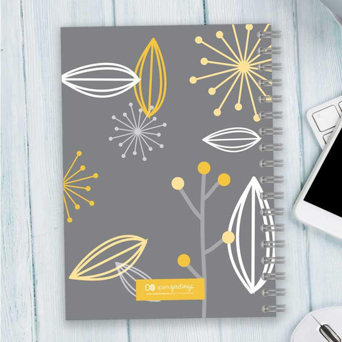 SparkGreetings® Deconstructed Nature Writing Journal, gray cover with yellow and white floral abstract design, spiral-bound notebook on a light wooden desk with a white smartphone and keyboard nearby.