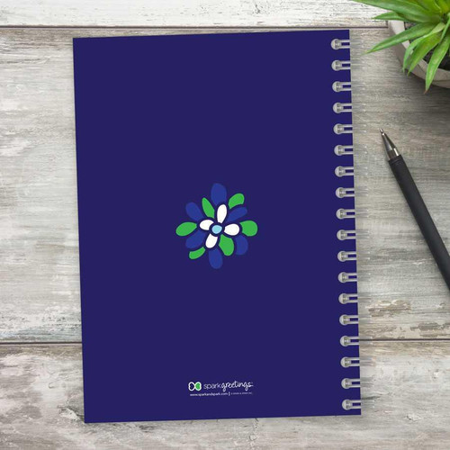 SparkGreetings® Stamped Blossoms Writing Journal, navy blue spiral-bound journal with a colorful floral design on the cover placed on a light wooden surface with a pen and green plant nearby