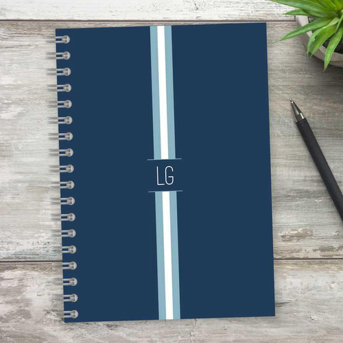 SparkGreetings® Elegant Ribbon Writing Journal, dark blue cover with light blue and white ribbon design, silver spiral binding, placed on a light wooden surface with a black pen and green plant nearby.