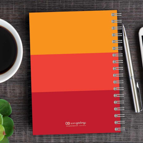 SparkGreetings® Shades of Color Writing Journal, vibrant orange and red spiral notebook placed on wooden desk with a cup of coffee, a pen, and a smartphone.
