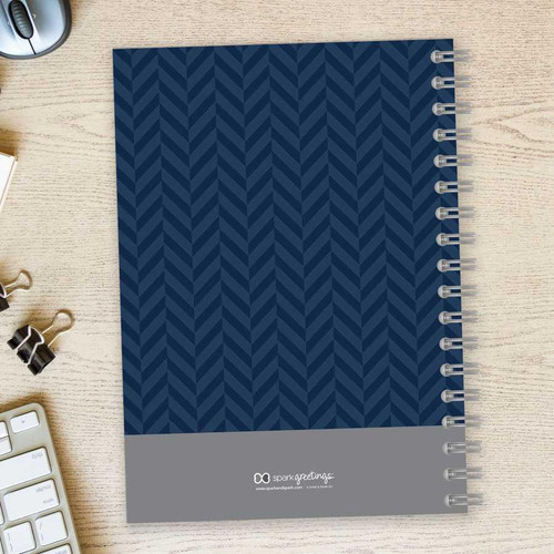 SparkGreetings® Herringbone Mood Writing Journal, navy blue herringbone pattern cover with gray section at the bottom, spiral binding on the right side, placed on a light wooden desk with a keyboard, paper clips, and a clock nearby.